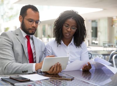 Focused coworkers checking reports on papers and on tablet. Business man and woman sitting in coffee shop, showing tablet and smartphone screens to each other. Teamwork concept